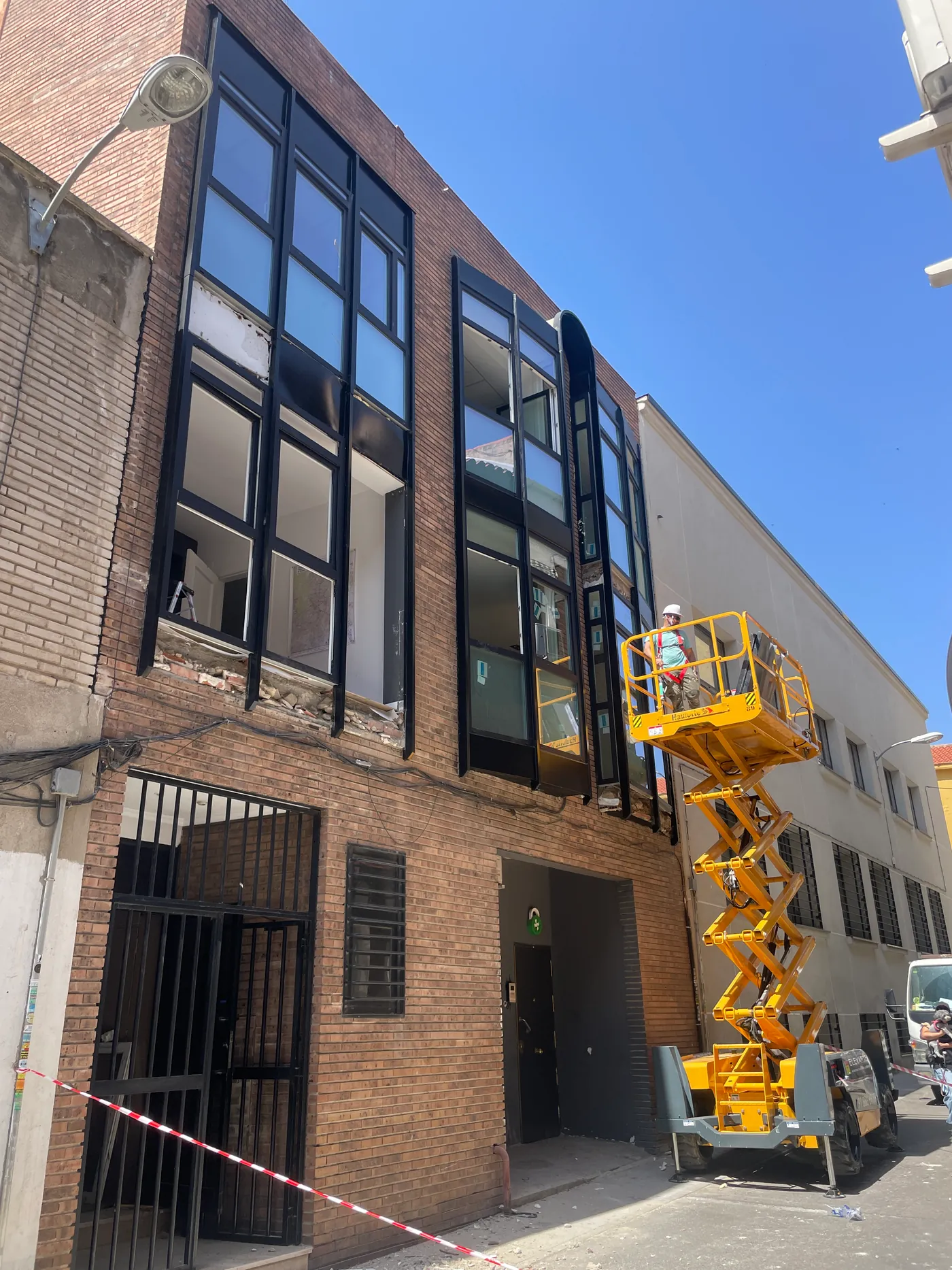 Fachada de ladrillo con miradores de perfil negro a dos alturas durante el montaje, con un operario en una plataforma elevadora de tijera