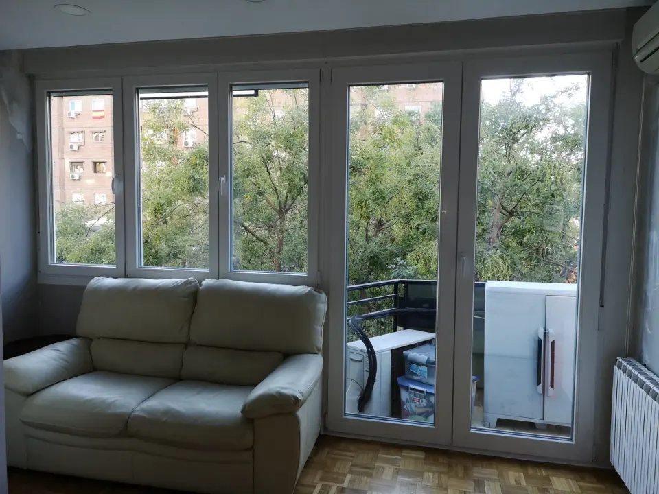 Salón con balconera y ventanas practicables de PVC blancas junto a un sofá, con vistas a árboles