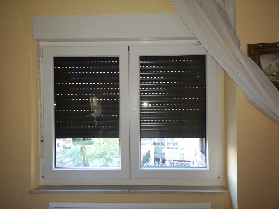 Ventana de PVC blanca de dos hojas con cajón de persiana encima, persiana oscura a media altura y alféizar interior de piedra