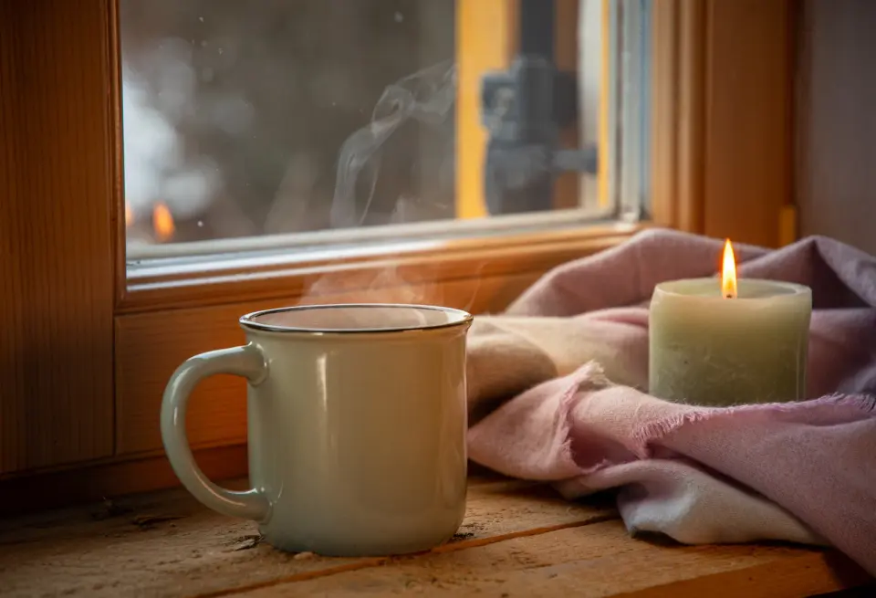 Vela encendida y taza humeante sobre el alféizar de madera de una ventana en invierno