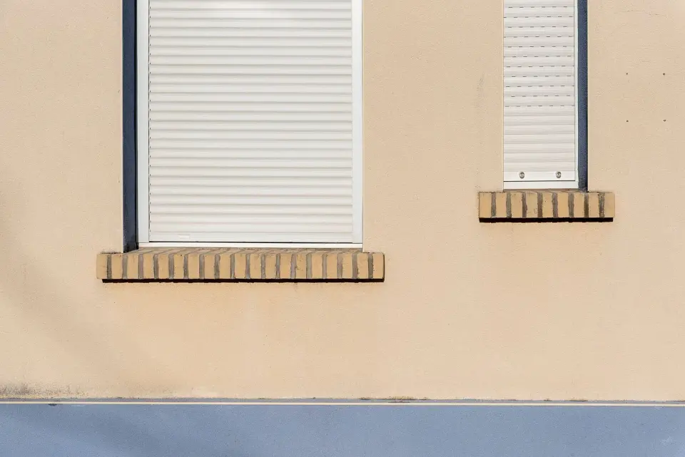 Fachada beige con dos ventanas y sus persianas blancas bajadas, una con las lamas cerradas y otra con las rendijas abiertas