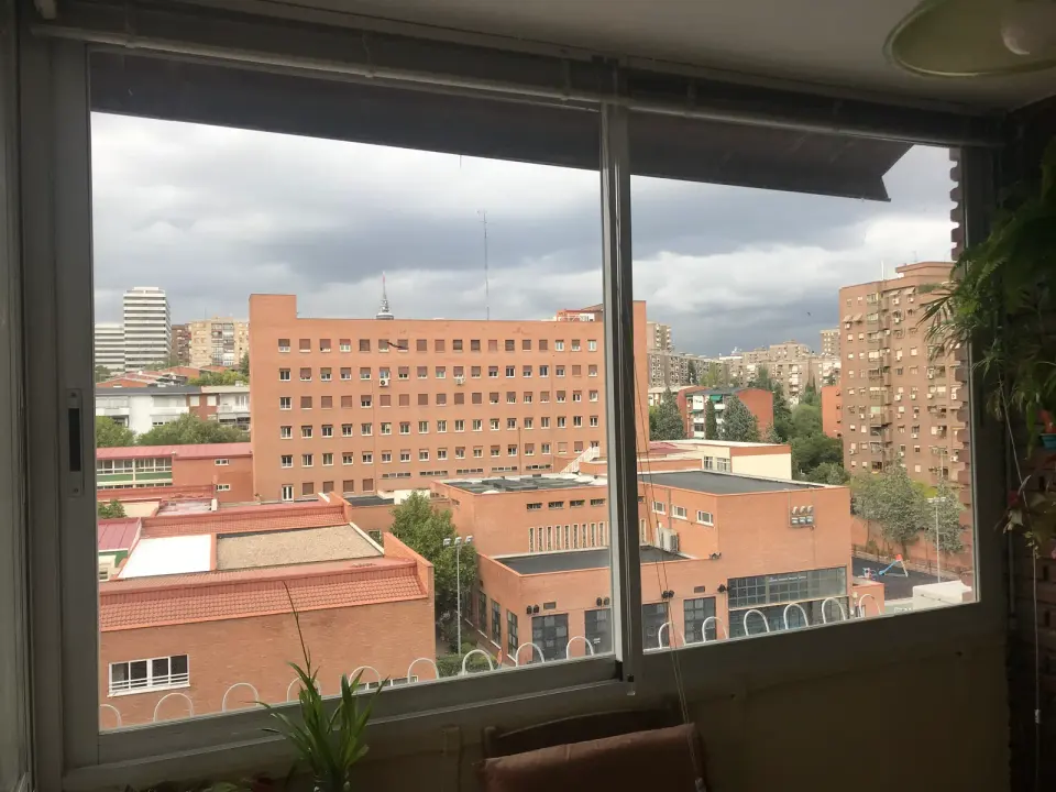 Ventana corredera antigua de aluminio de dos hojas vista desde dentro, con el cierre en el lateral y edificios de ladrillo al fondo