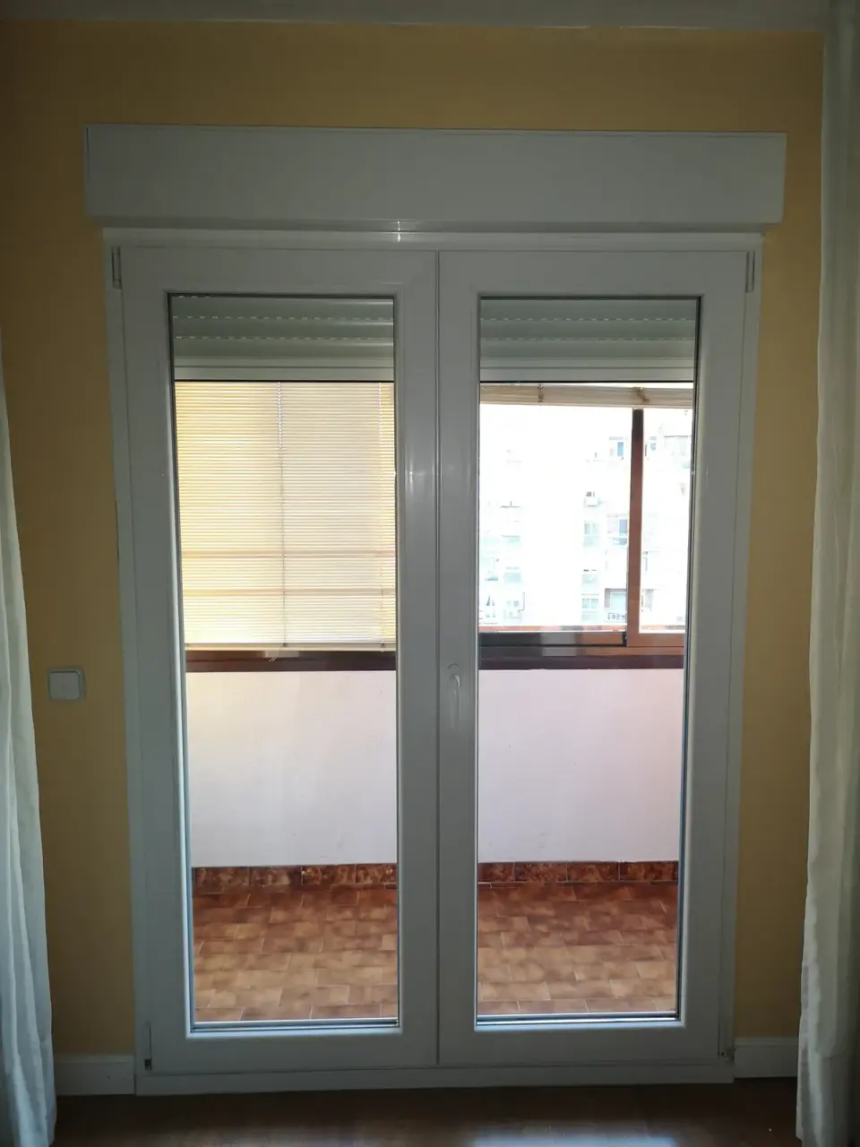Balconera de PVC blanca de dos hojas hasta el suelo con el cajón de persiana encima del marco, abierta a una terraza