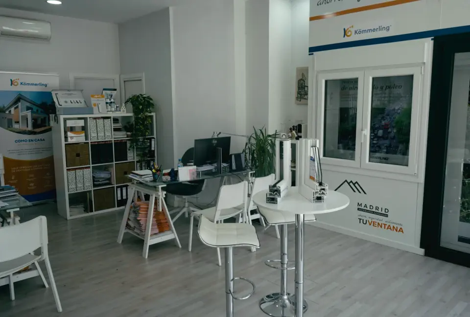 Tienda de Tu Ventana Madrid con muestras de ventanas en la pared bajo rótulos de Kömmerling, perfiles cortados sobre una mesa alta y la zona de oficina