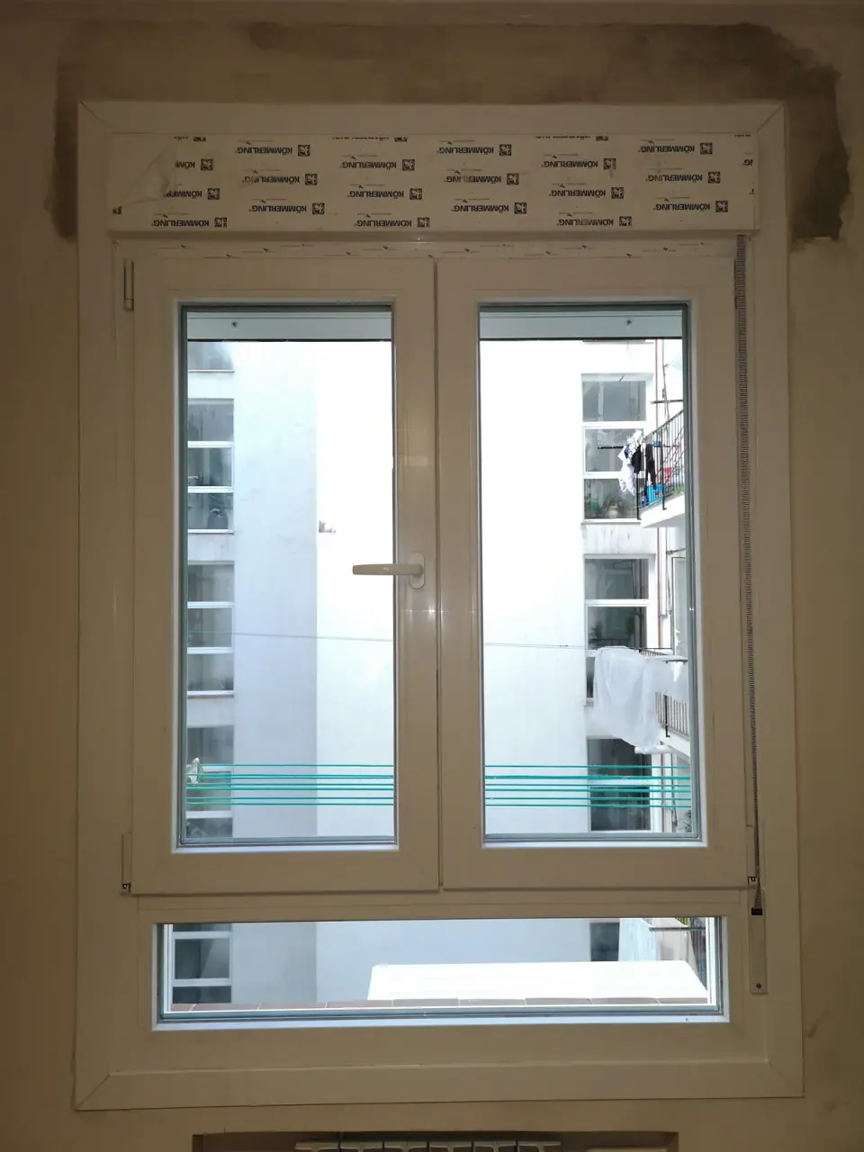 Ventana de PVC blanca de dos hojas recién colocada, con el cajón de persiana envuelto en un film protector con la marca Kömmerling impresa