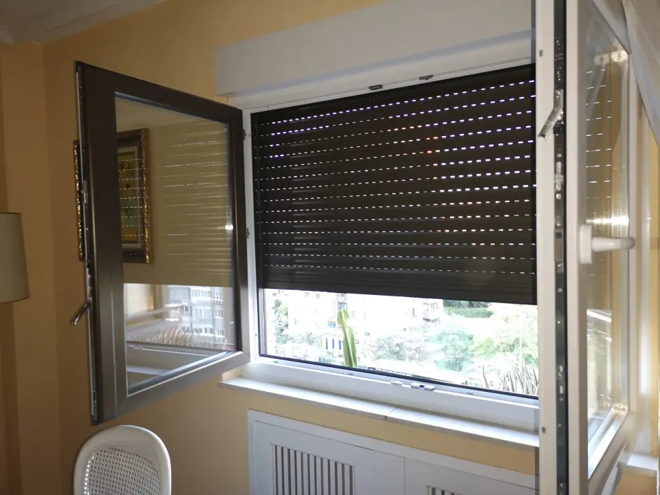 Ventana de PVC con una hoja abierta hacia dentro y la persiana de lamas oscuras a media altura bajo un cajón interior