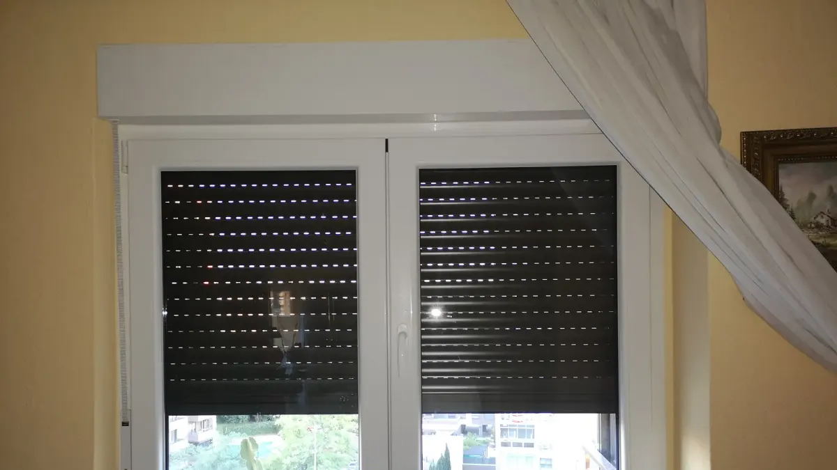 Ventana de PVC blanca de dos hojas con cajón de persiana encima, manilla central, persiana oscura a media altura y alféizar de piedra