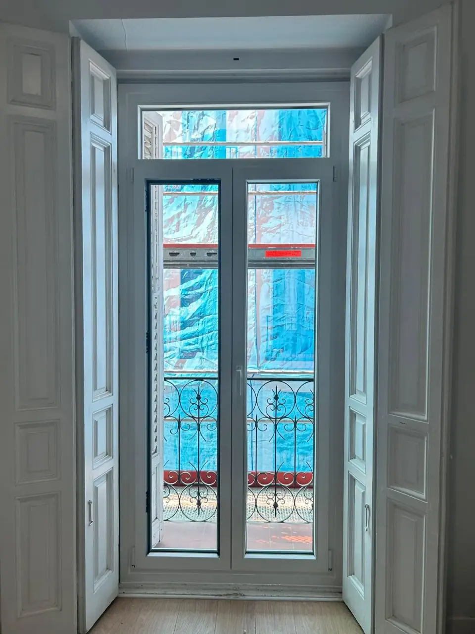 Balconera de PVC blanca de dos hojas con montante superior en un edificio antiguo, entre contraventanas interiores de madera abiertas