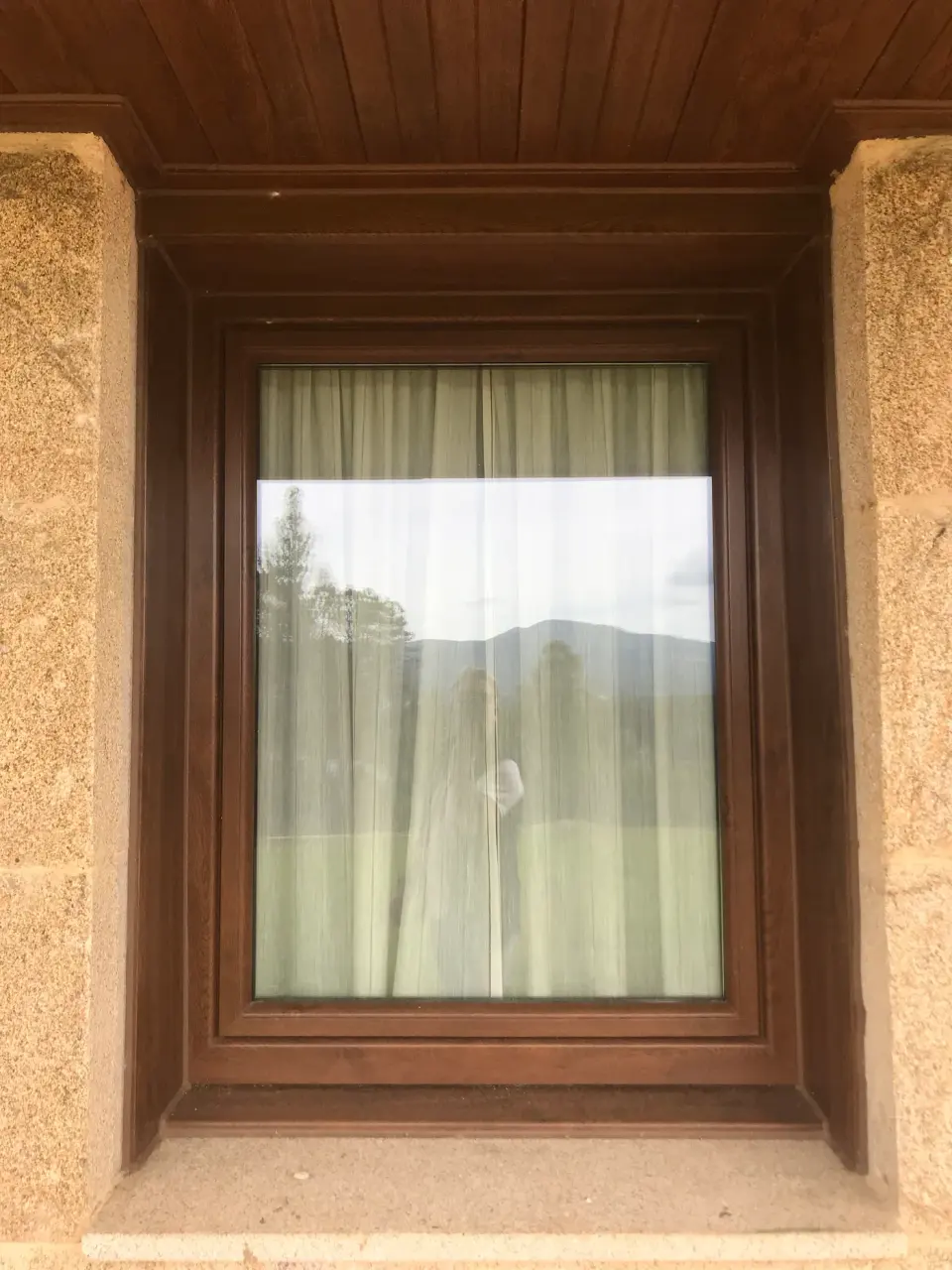 Ventana de PVC en color madera en un muro de piedra, con cortina blanca por dentro