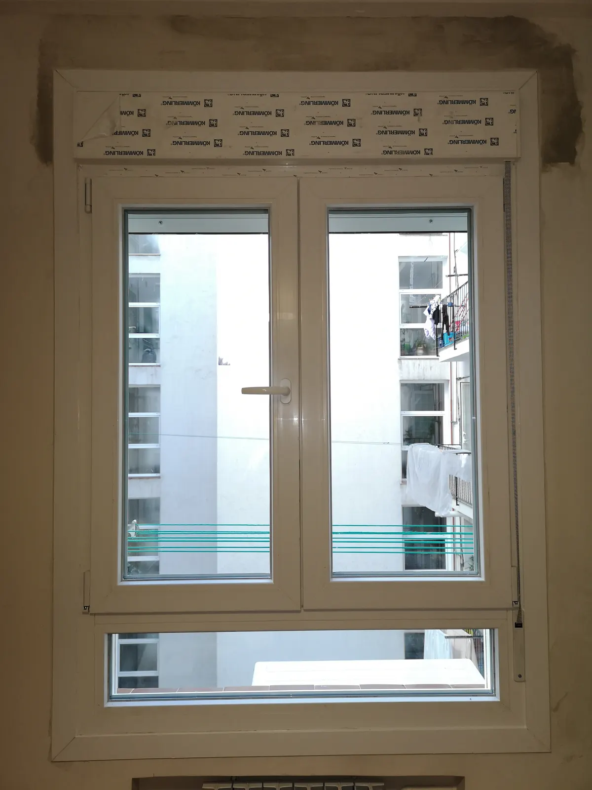 Ventana de PVC blanca de dos hojas recién instalada, con el cajón de persiana aún cubierto por el film protector de Kömmerling